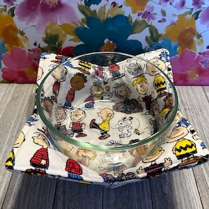 May include: A clear glass bowl sits in a square, white fabric bowl cozy. The cozy is patterned with cartoon characters in various poses. The background is a floral pattern.