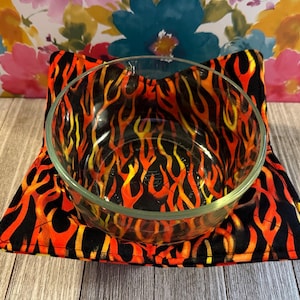 May include: A clear glass bowl sits inside a microwave bowl cozy. The cozy is black with a vibrant flame pattern in shades of orange and yellow. The cozy is square with a triangular shape.