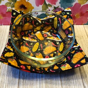 May include: A black fabric bowl cozy with a taco, corn, and chili pepper print. The cozy is holding a clear glass bowl.
