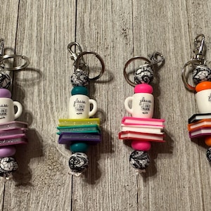 May include: Four keychains with silver rings, each featuring a white mug with the text "please read again", stacked books, and decorative beads in various colors like pink, purple, green, and orange. The beads have a black and white lace pattern.