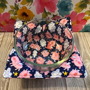 May include: A blue and white fabric bowl cozy with a pink pig design. The cozy is filled with a clear glass bowl.