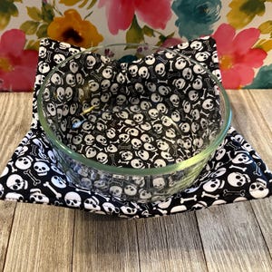 May include: A clear glass bowl sits inside a black and white skull-patterned bowl cozy. The cozy is square-shaped with a black background and white skulls and crossbones. The bowl is on a wooden surface.
