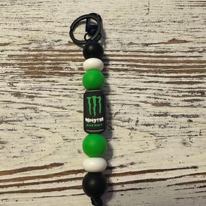 K&ouml;nnte beinhalten: Ein Schl&uuml;sselanh&auml;nger mit einem schwarzen Metallring, der an einer Reihe runder Silikonperlen in Schwarz, Wei&szlig; und Gr&uuml;n befestigt ist. Eine zentrale zylindrische Perle zeigt das Monster Energy Logo in gr&uuml;nem und wei&szlig;em Text auf schwarzem Grund.
