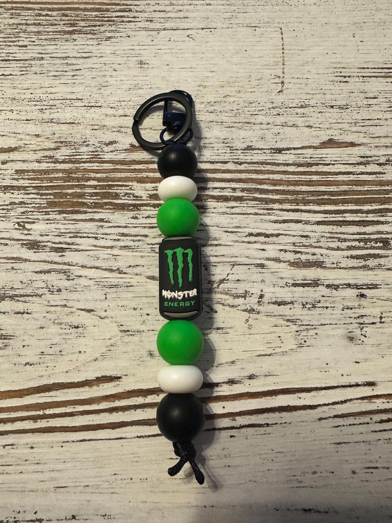 K&ouml;nnte beinhalten: Ein Schl&uuml;sselanh&auml;nger mit einem schwarzen Metallring, der an einer Reihe runder Silikonperlen in Schwarz, Wei&szlig; und Gr&uuml;n befestigt ist. Eine zentrale zylindrische Perle zeigt das Monster Energy Logo in gr&uuml;nem und wei&szlig;em Text auf schwarzem Grund.