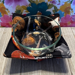 May include: A black bowl cozy with a guitar print, holding a clear glass bowl. The guitar pattern features various brown, white, and black electric guitars. The cozy is square with folded corners.