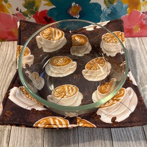 May include: A brown quilted hot pad with a coffee cup design. The hot pad is made of fabric and has a quilted texture. The coffee cups are filled with a latte design. The hot pad is perfect for protecting surfaces from hot dishes.