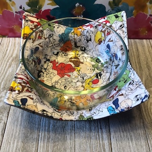 May include: A white bowl cozy with a colorful cartoon print of various characters. The cozy is made of fabric and has a square shape. A clear glass bowl is sitting inside the cozy.