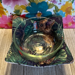May include: A clear glass bowl sits inside a square, quilted bowl cozy. The cozy features a dark green and black tropical leaf pattern with a dinosaur design. The cozy has a white border and is on a wooden surface.