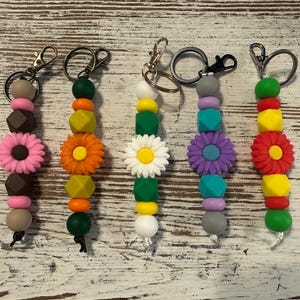May include: Five colorful beaded keychains with flower and geometric designs. Each keychain features a variety of beads in shades of pink, orange, yellow, green, and purple, with a metal clasp for attaching to keys or bags.