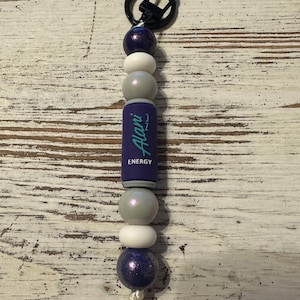 K&ouml;nnte beinhalten: Ein Schl&uuml;sselanh&auml;nger mit einer vertikalen Anordnung von violetten, wei&szlig;en und silbernen Perlen, gekr&ouml;nt von einem schwarzen Metallring. Eine zentrale zylindrische violette Perle zeigt den Schriftzug Alani Nu ENERGY in t&uuml;rkiser und wei&szlig;er Schrift.