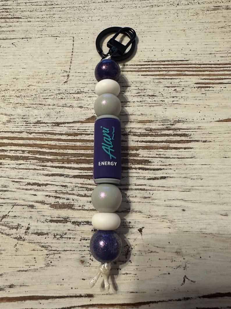 K&ouml;nnte beinhalten: Ein Schl&uuml;sselanh&auml;nger mit einer vertikalen Anordnung von violetten, wei&szlig;en und silbernen Perlen, gekr&ouml;nt von einem schwarzen Metallring. Eine zentrale zylindrische violette Perle zeigt den Schriftzug Alani Nu ENERGY in t&uuml;rkiser und wei&szlig;er Schrift.