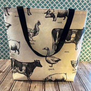 May include: Cream-colored tote bag featuring black and white illustrations of farm animals, including cows, pigs, chickens, and a goose. The bag has black handles and the animals are labeled with their sounds. A charming and practical accessory.