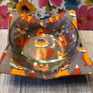May include: A gray and orange fabric bowl cozy with a pattern of foxes wearing glasses and hats. The cozy is holding a clear glass bowl.