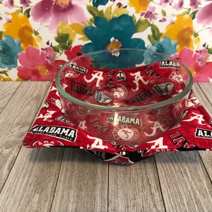 May include: Red and black bowl cozy with the University of Alabama logo and the word "Alabama" repeated throughout. A clear glass bowl is sitting on top of the cozy.