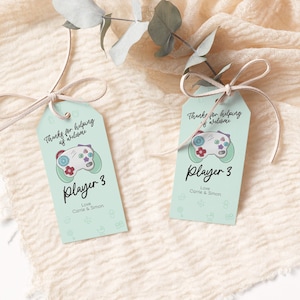 May include: Two light blue favor tags with a white and green video game controller graphic. The text on the tags reads "Thanks for helping us welcome Player 3 Love Carrie & Simon".