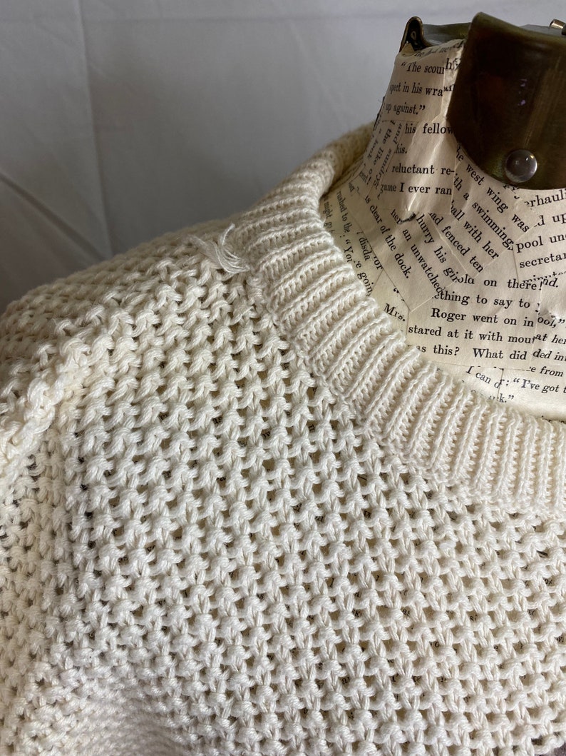 May include: A cream-colored knitted sweater with a loose, open weave. The sweater is on a mannequin and has a crew neck.