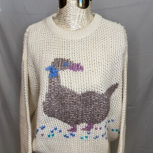 May include: A cream-colored knitted sweater with a brown, purple, and blue embroidered goose design. The goose is walking on a trail of blue, purple, and teal hearts.