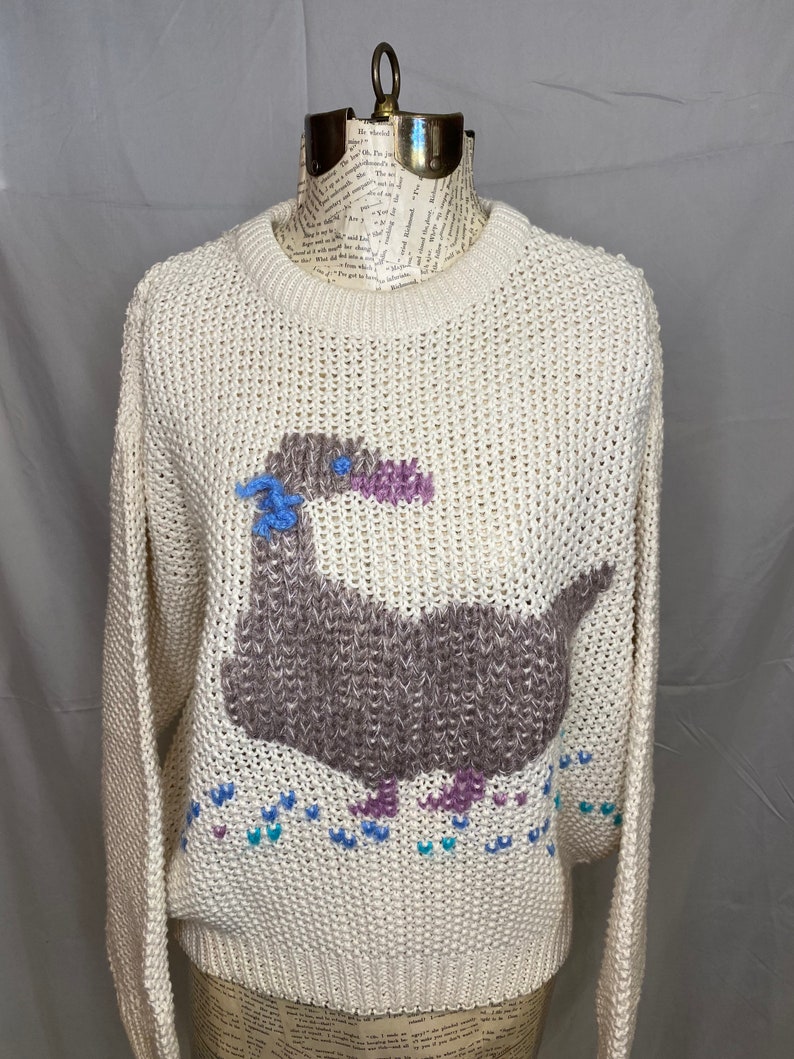 May include: A cream-colored knitted sweater with a brown, purple, and blue embroidered goose design. The goose is walking on a trail of blue, purple, and teal hearts.