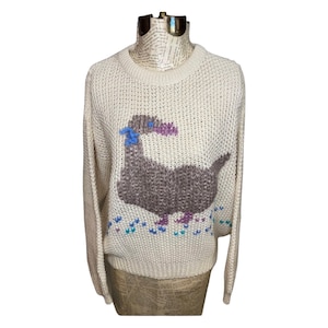 May include: A cream-colored knitted sweater with a brown and purple dinosaur design. The dinosaur has blue eyes and a pink beak. The sweater has a crew neck and long sleeves.
