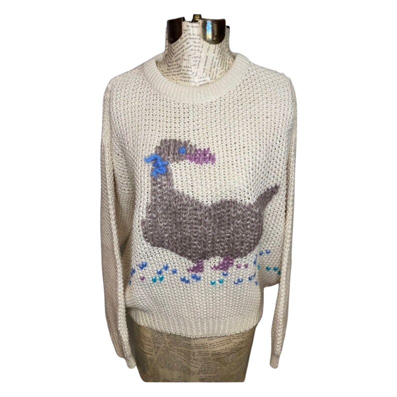 May include: A cream-colored knitted sweater with a brown and purple dinosaur design. The dinosaur has blue eyes and a pink beak. The sweater has a crew neck and long sleeves.