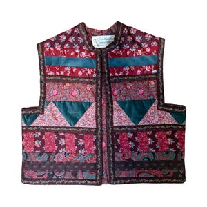 May include: A quilted vest with a floral pattern in shades of burgundy, pink, and teal. The vest features teal velvet accents and a brown trim. The vest has a high collar and a button closure.