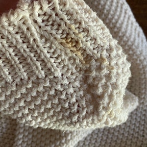 May include: Close-up of a cream-colored knitted sweater with a small yellow stain.