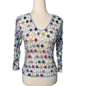 May include: A colorful crochet top with a V-neckline. The long-sleeved top features a light blue trim and is adorned with various colored floral patterns, including red, yellow, green, and purple. The open weave design is accented with small beads.