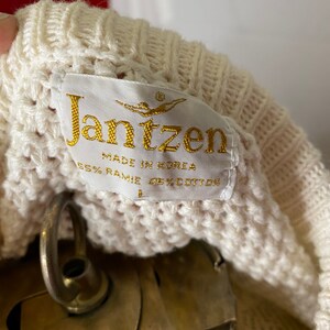 May include: A white knitted sweater with a cream-colored label that reads "Jantzen" in gold lettering. The label also says "Made in Korea" and "65% Ramie 35% Cotton".