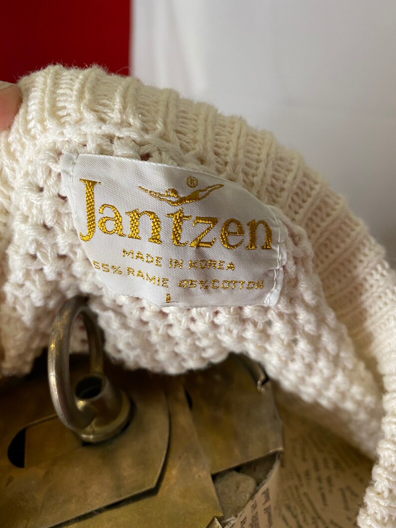 May include: A white knitted sweater with a cream-colored label that reads "Jantzen" in gold lettering. The label also says "Made in Korea" and "65% Ramie 35% Cotton".
