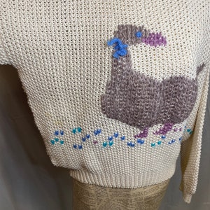 May include: A cream-colored knitted sweater with a brown and purple bird design. The bird has a blue flower on its head and is surrounded by blue, purple, and white hearts.
