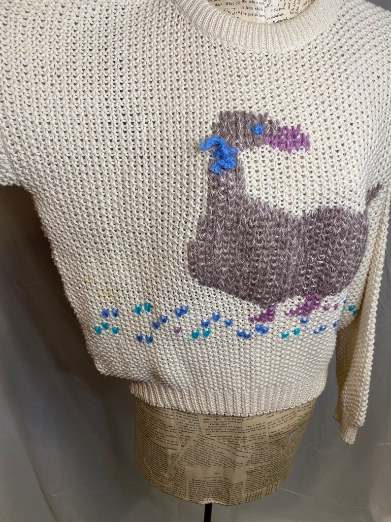 May include: A cream-colored knitted sweater with a brown and purple bird design. The bird has a blue flower on its head and is surrounded by blue, purple, and white hearts.