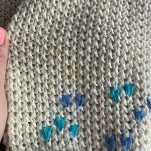 May include: Close-up of a cream-colored knitted fabric with a subtle pattern of blue and turquoise embroidered flowers.