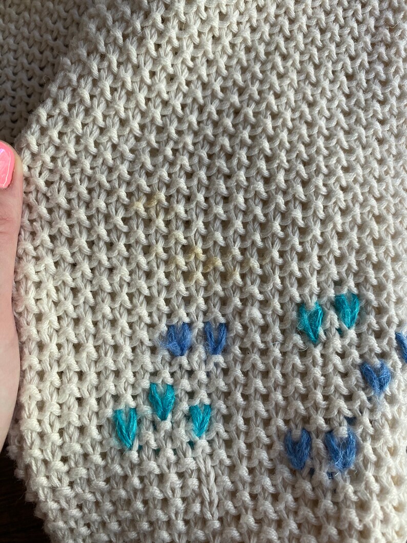 May include: Close-up of a cream-colored knitted fabric with a subtle pattern of blue and turquoise embroidered flowers.