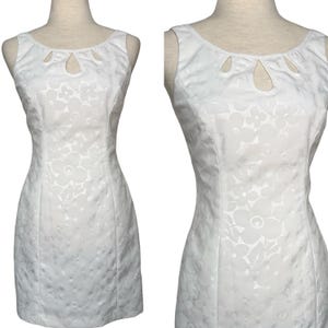 May include: White sleeveless dress with a floral pattern and keyhole neckline. The dress is fitted and appears to be made of a textured fabric. The dress is displayed on a mannequin.