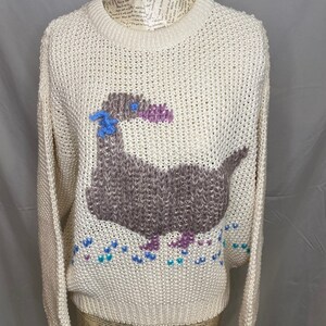 May include: A cream-colored knitted sweater with a brown, purple, and blue embroidered duck design. The duck is surrounded by small blue, purple, and white hearts.