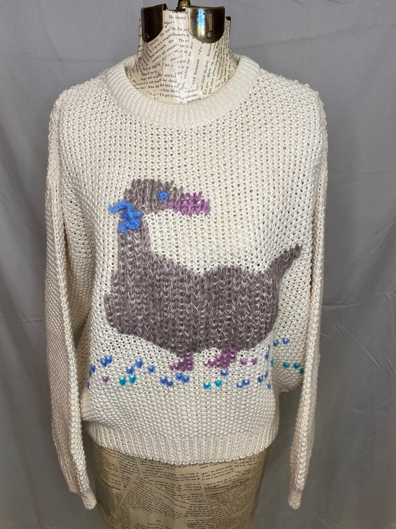 May include: A cream-colored knitted sweater with a brown, purple, and blue embroidered duck design. The duck is surrounded by small blue, purple, and white hearts.