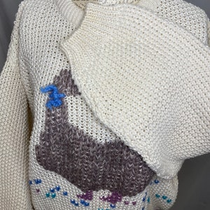 May include: A white knitted sweater with a brown llama design. The llama has blue and purple accents. The sweater has a loose fit and a crew neck.