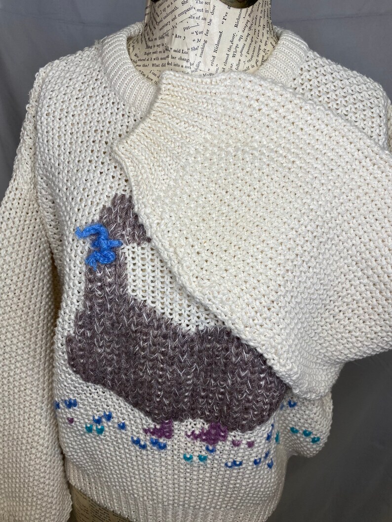May include: A white knitted sweater with a brown llama design. The llama has blue and purple accents. The sweater has a loose fit and a crew neck.