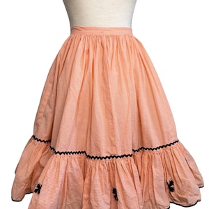 May include: A peach and white gingham skirt with a tiered ruffle hem. The skirt has black trim and decorative black floral accents. The skirt is displayed on a mannequin.
