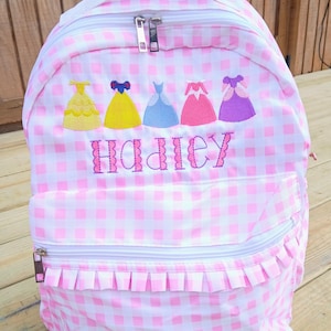 May include: Pink and white gingham backpack with a white handle and zipper. The backpack features embroidered princess dresses in yellow, blue, pink, and purple, with the name "Hadley" embroidered below. A front pocket and ruffled trim complete the design.