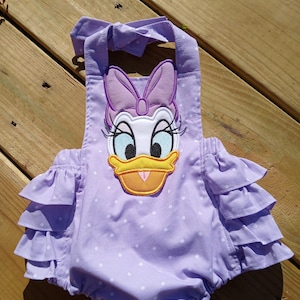 May include: A lavender romper with white polka dots and ruffled sleeves. The romper features an embroidered Daisy Duck face with a yellow beak, black eyelashes, and a purple bow. The straps tie at the shoulders.