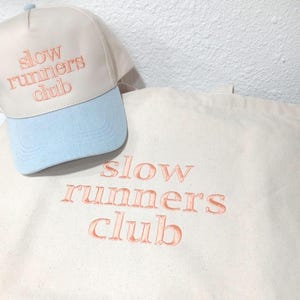 May include: A cream-colored baseball cap with a light blue brim and a cream-colored canvas tote bag. Both items feature the text "slow runners club" embroidered in a peach-colored thread. The cap has a structured front panel and a button on top.