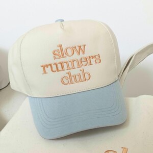 May include: A baseball cap featuring a cream-colored crown and a light blue brim. The front of the crown displays the embroidered text "slow runners club" in a copper-colored thread. The cap has a structured front panel and a button on top.