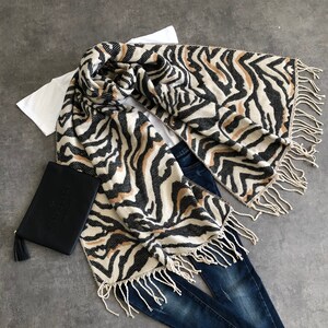 May include: A patterned scarf with a zebra design in black, tan, and cream, featuring fringed edges. A black clutch bag with a tassel is also visible. The scarf is styled with a white t-shirt and blue jeans.