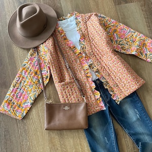 May include: A tan fedora hat, a floral print jacket, a white t-shirt, a brown Coach crossbody bag, and blue jeans are arranged on a wooden surface. The jacket has a pink, yellow, and orange floral pattern.