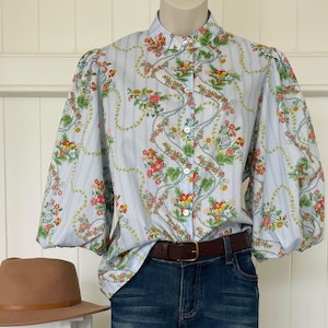 May include: A light blue floral print blouse with vertical stripes and puff sleeves. The blouse has a high collar and button-down front. A brown belt and blue jeans are also visible. A brown hat is in the foreground.