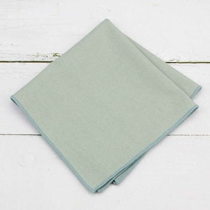 May include: A folded, square pocket square in a pale green hue with a contrasting blue border. The pocket square is made of a textured fabric and is displayed on a white wooden surface.