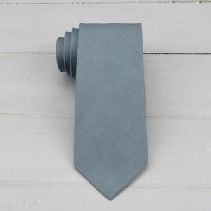 May include: A solid slate blue necktie with a visible woven linen texture. The tie is folded at the top, showing the layered fabric construction, and tapers to a pointed tip at the bottom, displaying a consistent matte finish throughout the piece.