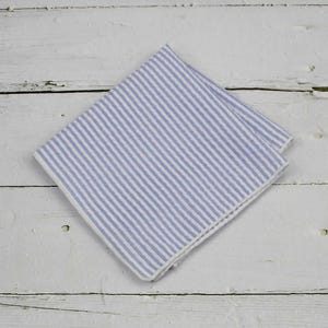 May include: A folded blue and white striped handkerchief. The fabric has vertical stripes of light blue and white. The handkerchief is made of a soft, likely linen, material. The background is a white wooden surface.