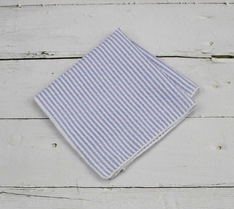 May include: A folded blue and white striped handkerchief. The fabric has vertical stripes of light blue and white. The handkerchief is made of a soft, likely linen, material. The background is a white wooden surface.